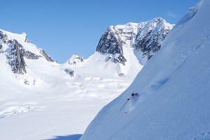 A skier descends a steep slope in the Alaska Range, skiing powder, with large mountains in the background. 